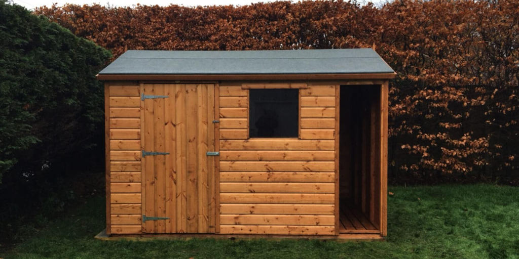 Wooden garden shed with window and sloped roof surrounded by green and brown foliage